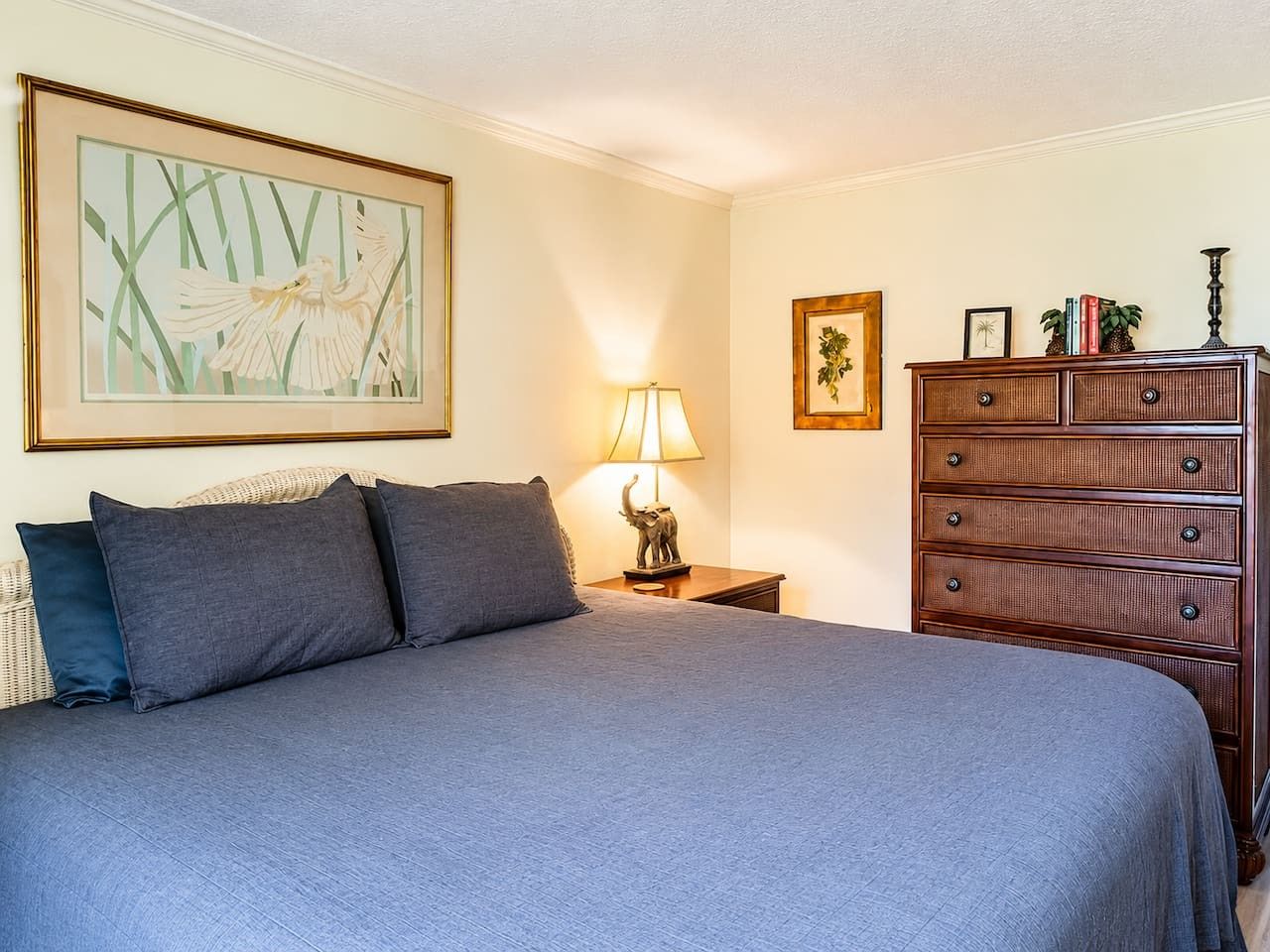 Un dormitorio con cama, almohadas, mesita de noche con lámpara, cómoda y decoración en la pared.
