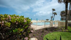 A pool area with a fence, palm trees, and a building in the background. - Coquina B101 Ground-Floor Coastal Condo (Saint Augustine)