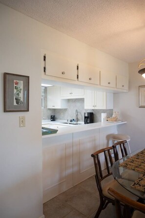 A compact kitchen with white cabinets, a coffee maker, and a dining area with a glass table and wooden chairs. - Coquina B101 Ground-Floor Coastal Condo (Saint Augustine)
