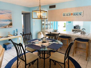 Un comedor con mesa de vidrio, sillas y una araña. Hay una zona de cocina con microondas y hervidor. La habitación tiene una decoración azul y un letrero que dice 'BEACH HOUSE'.