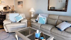 Ein Wohnzimmer mit einer Couch, einem Sessel, einem Couchtisch und einem Essbereich im Hintergrund.