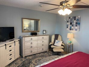 A bedroom with a bed, a dresser, a chair, a TV, a ceiling fan, and a wall clock. - Ground-Floor Oceanfront Condo | Steps to Beach (Saint Augustine)