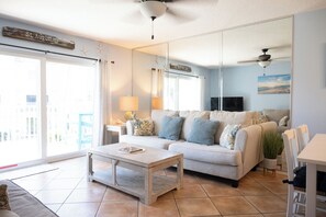 A living room with a sofa, coffee table, and a TV. There is a large window with curtains, a ceiling fan, and a sign on the wall.