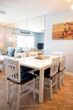 A dining area with a white table and chairs, a painting on the wall, and a view into the living room.