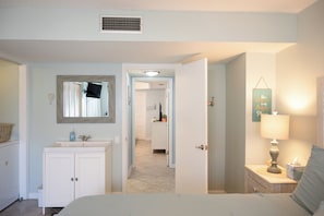 A bedroom with a bed, a bedside table with a lamp, a nightstand, a dresser, a mirror, and a door leading to a hallway.