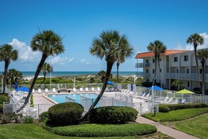 A pool area with lounge chairs and umbrellas, palm trees, and a building with balconies.