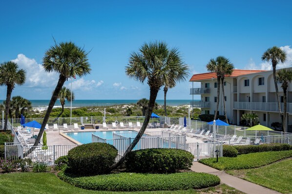 A pool area with lounge chairs and umbrellas, palm trees, and a building with balconies.