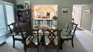 A dining room with a round table and six chairs, a kitchen in the background, and a hallway leading to a door. - The Pearl by Sandpiper | Near Beach (Seabrook Island)