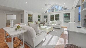 A spacious living room with white furniture, a ceiling fan, large windows, and a bookshelf. - Mystic Pines by Sandpiper | Near Beach (Seabrook Island)