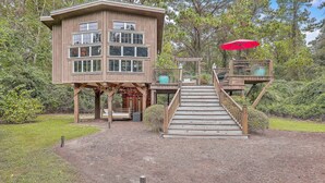 Ein hölzernes Baumhaus mit mehreren Ebenen, einem roten Sonnenschirm und einer Holztreppe, die dorthin führt.
