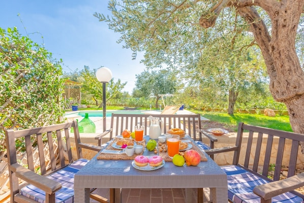 An outdoor dining area with a table set for breakfast, surrounded by wooden chairs and a view of a swimming pool.