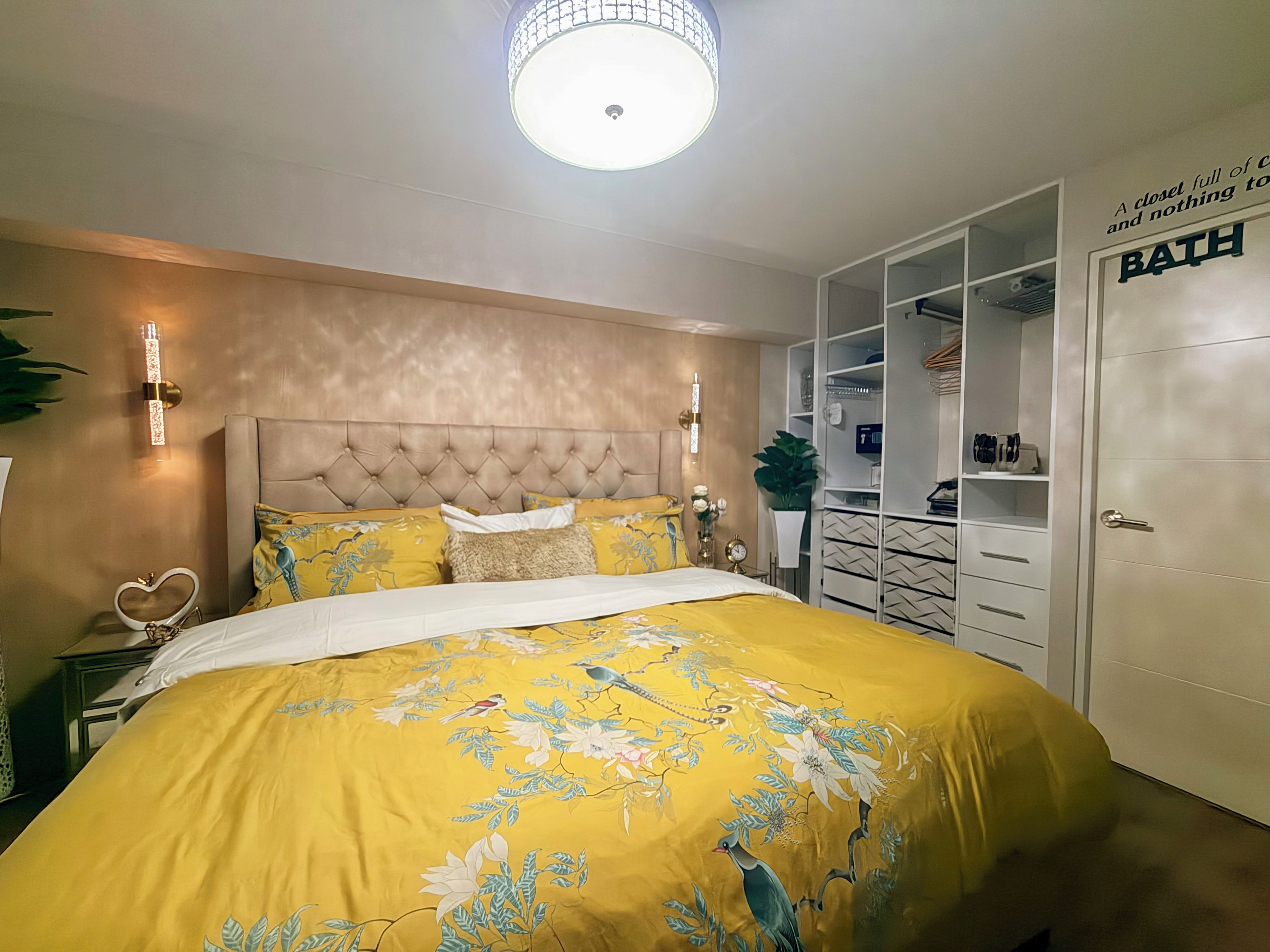 Une chambre à coucher avec une tête de lit capitonnée, un lit recouvert d’une literie à motifs floraux jaunes, des lampes de chevet et une armoire sur laquelle est apposé un panneau indiquant « SALLE DE BAIN ».