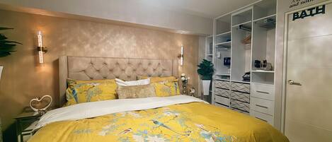 A bedroom with a tufted headboard, a bed with yellow floral bedding, bedside lamps, and a wardrobe with a 'BATH' sign.