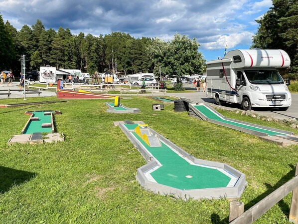 Un parcours de minigolf avec un terrain verdoyant, un camping-car blanc et une forêt en arrière-plan.
