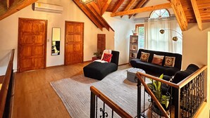 Un loft spacieux avec des poutres apparentes, un canapé noir, un escalier en bois et une grande fenêtre.