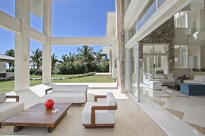 A spacious indoor area with a large glass wall offering a view of a well-maintained lawn and palm trees. There are white sofas and a wooden coffee table with a red vase.