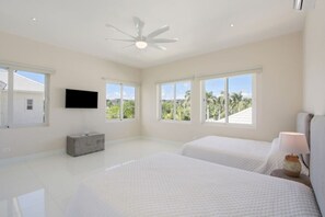A spacious bedroom with two beds, a ceiling fan, a TV, and large windows offering a view of palm trees.