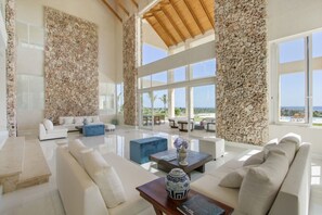 A spacious living room with a white sofa set, a blue ottoman, and a coffee table with a decorative vase. Large windows offer a view of the ocean.