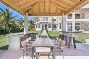 An outdoor patio with a wooden pergola, a long dining table with chairs, and a view of a modern building with a stone facade and large windows.