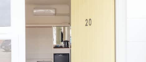 A hotel room with a yellow door marked '20', a white wardrobe, a small kitchenette with a coffee maker, and a mirror above the counter.