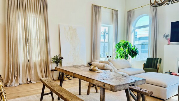 Una sala de estar acogedora con una mesa de madera, un banco y un sofá. Hay una ventana grande con cortinas, una planta verde y una silla.