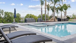 Un área de piscina con sillones de descanso, una mesa y un espacio de estar con vista a palmeras y un cielo despejado.