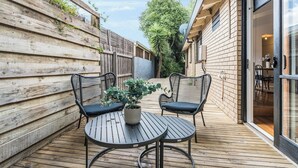 Une terrasse en bois avec deux chaises en osier noir et une table, une plante en pot et une porte vitrée donnant sur une pièce.