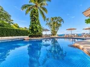 A clear blue pool with a tiled edge, surrounded by lounge chairs and umbrellas, with palm trees and a hedge in the background.