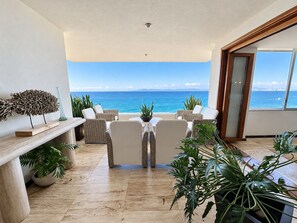 A sunlit balcony with a view of the sea, wicker furniture, and potted plants.