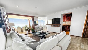 A modern living room with a large white sofa, a coffee table, and a TV mounted on the wall. There is a view of the ocean through large windows. - Beachfront Oasis with a View - Puerto Vallarta (Puerto Vallarta)