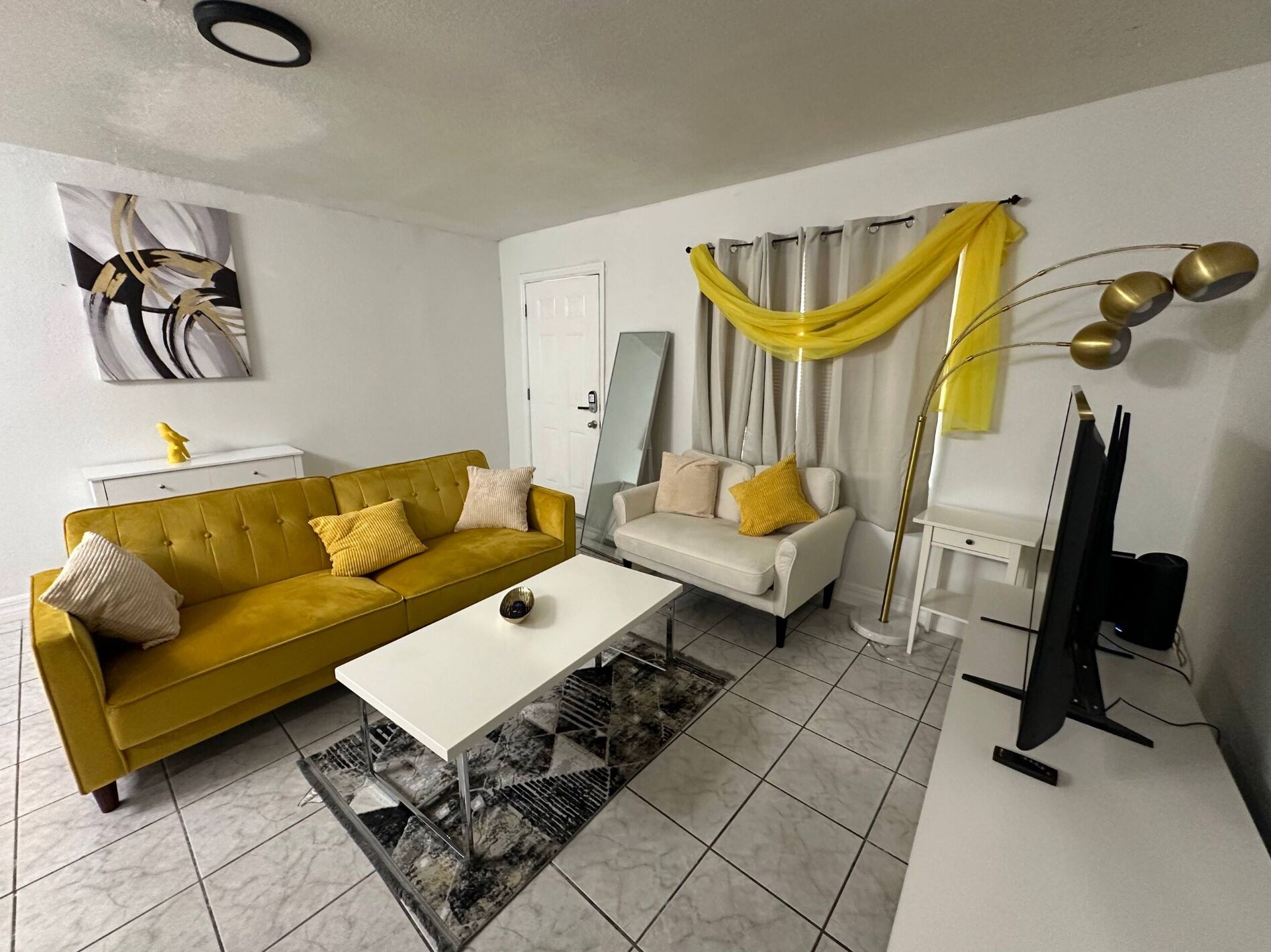 Ein Wohnzimmer mit gelbem Sofa, weißem Sofa, weißem Couchtisch, Stehlampe und Flachbildfernseher.