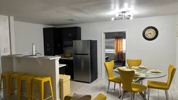 A compact kitchen with dark cabinets, a stainless steel refrigerator, and a dining area with yellow chairs and a glass table.