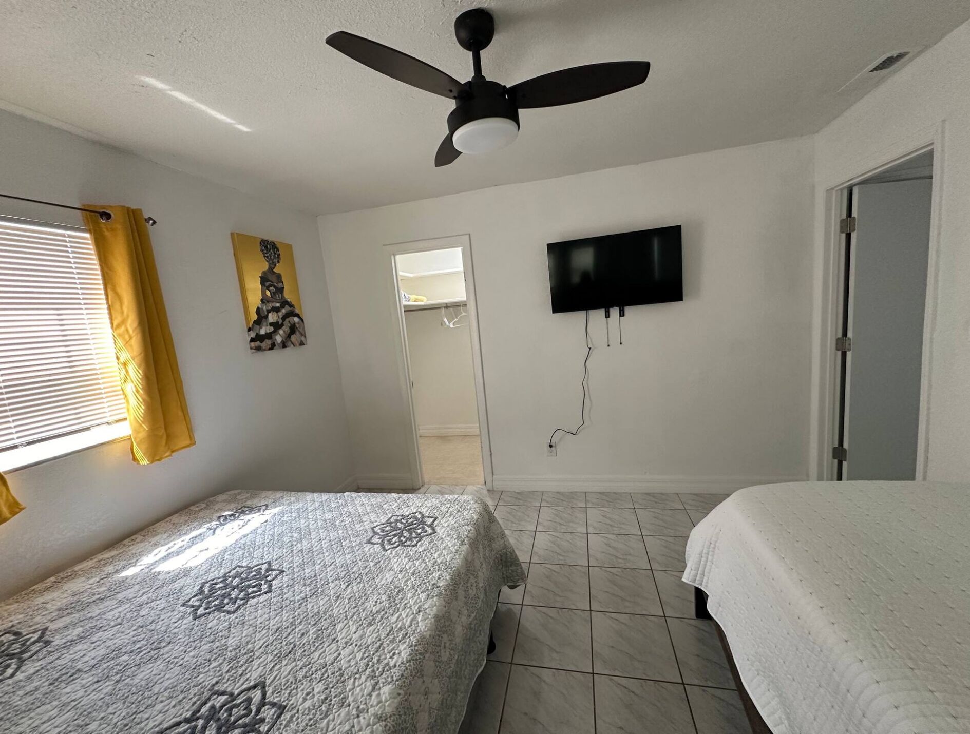 A bedroom with two beds, a ceiling fan, a TV mounted on the wall, and a window with curtains.