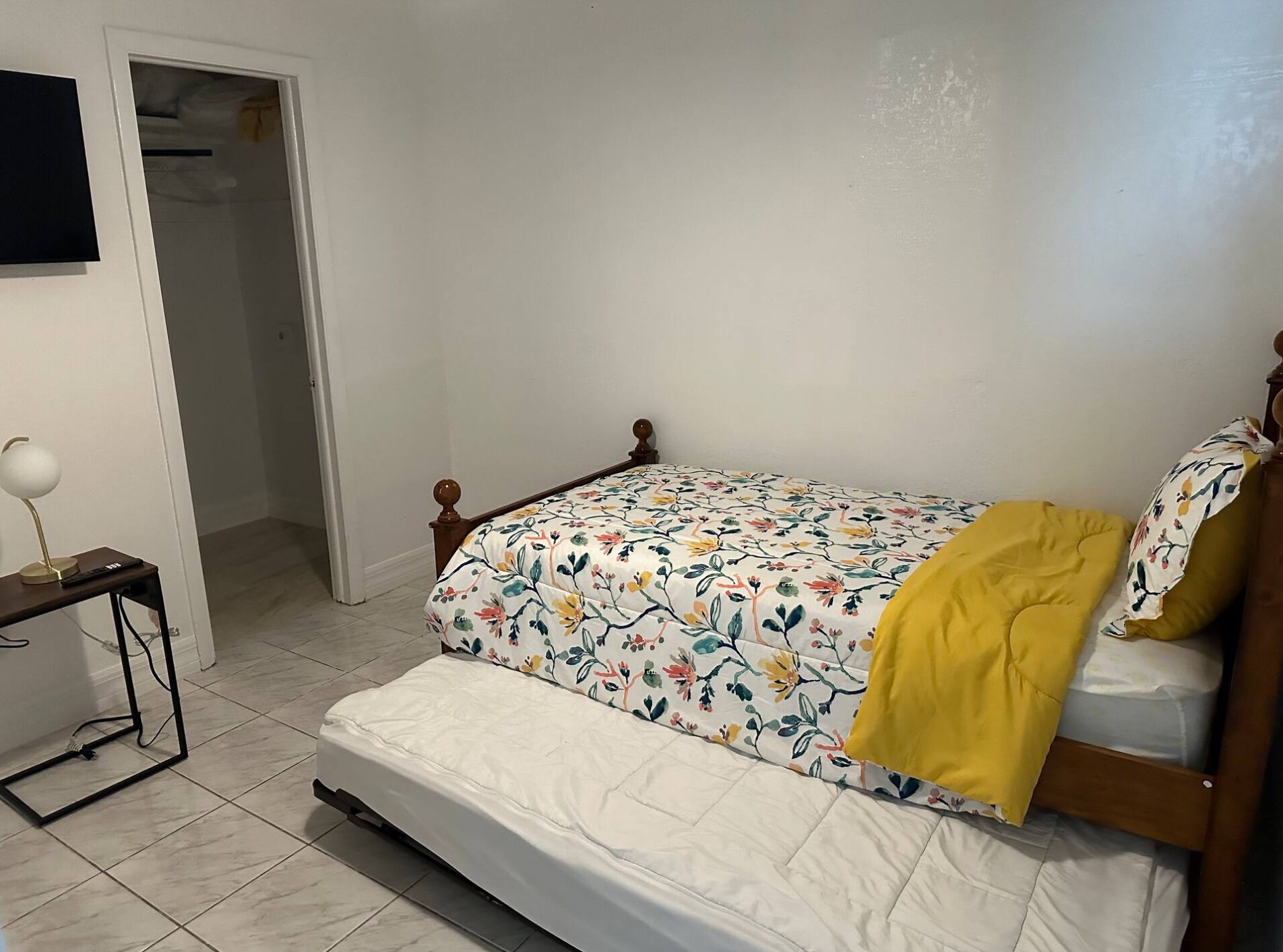 A small, neatly arranged bedroom with a single bed, a bedside table with a lamp, and a door leading to another room.