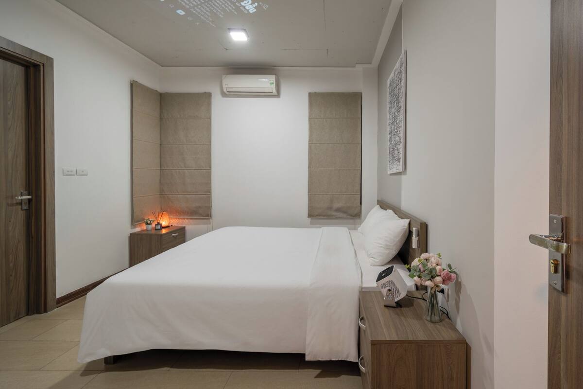 A modern bedroom with a large bed, a bedside table with a lamp, an air conditioner, and a wall-mounted projector.