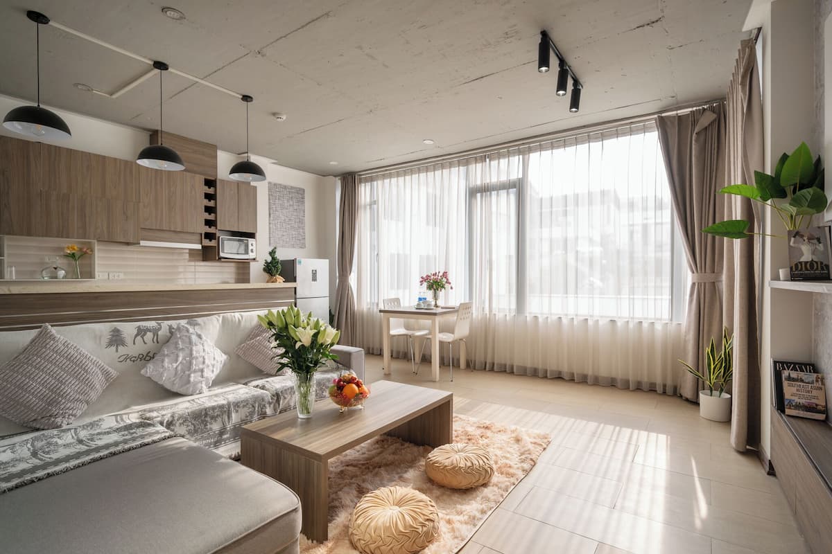 A modern living room with a sofa, coffee table, and dining area. There is a kitchen with wooden cabinets and a microwave. The room has large windows with curtains and a view of the city.