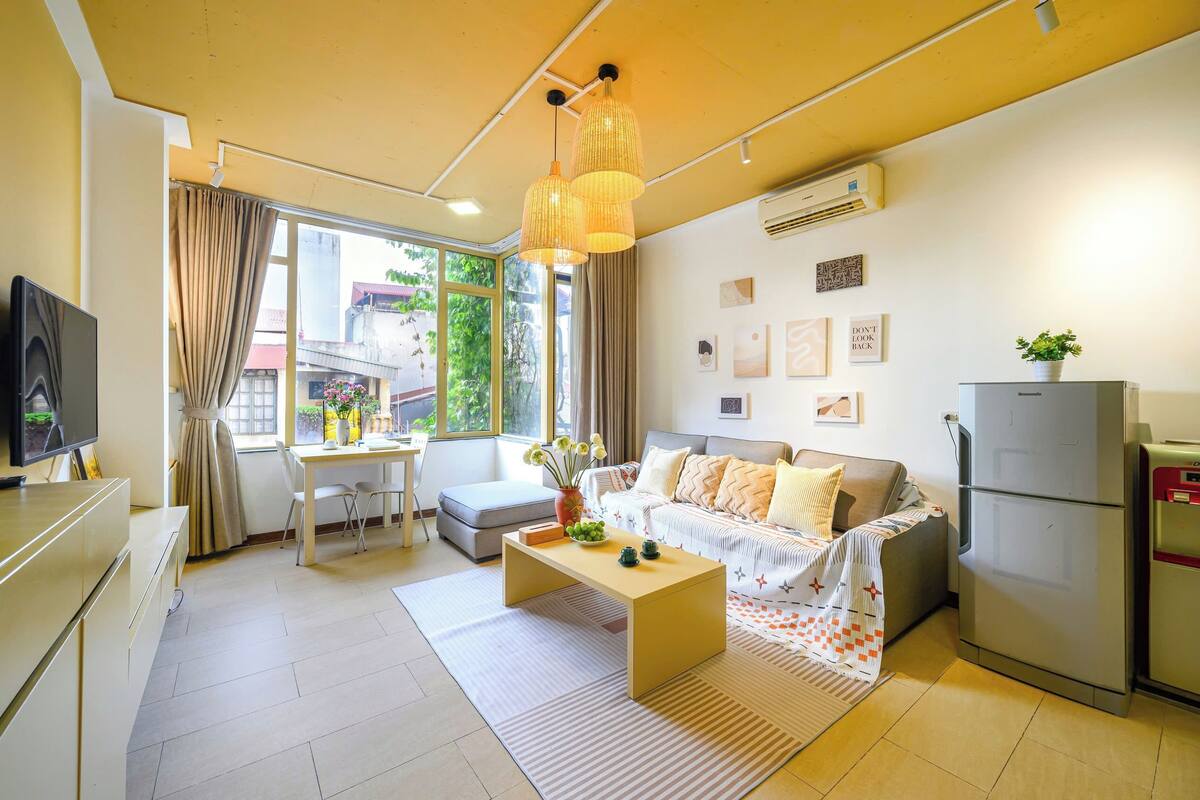 A modern living room with a sofa, coffee table, and a TV. There is a dining area with a table and chairs. The room has large windows with curtains.
