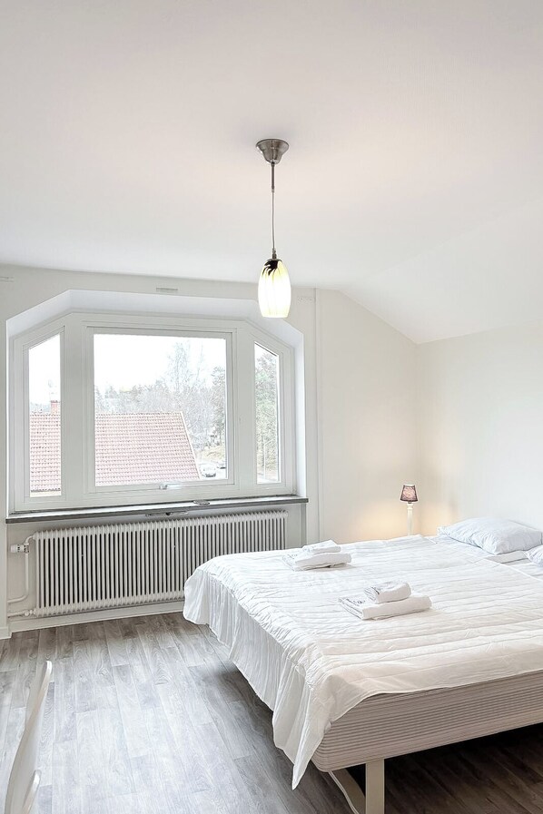 Un dormitorio con un ventanal grande, un radiador, una cama con ropa de cama blanca y una lámpara colgante.