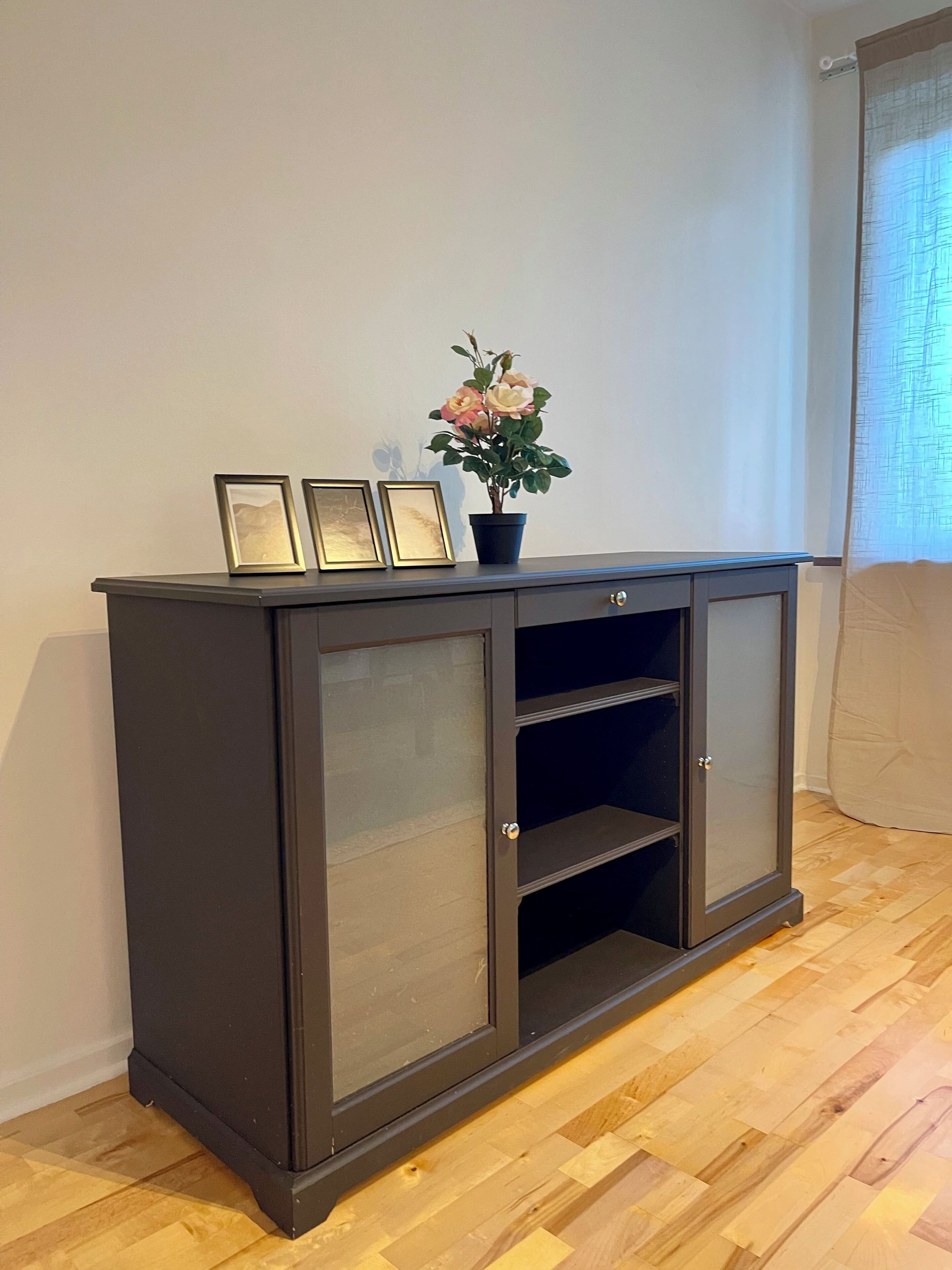 A dark wooden cabinet with glass doors and shelves, placed against a white wall with a window on the right.