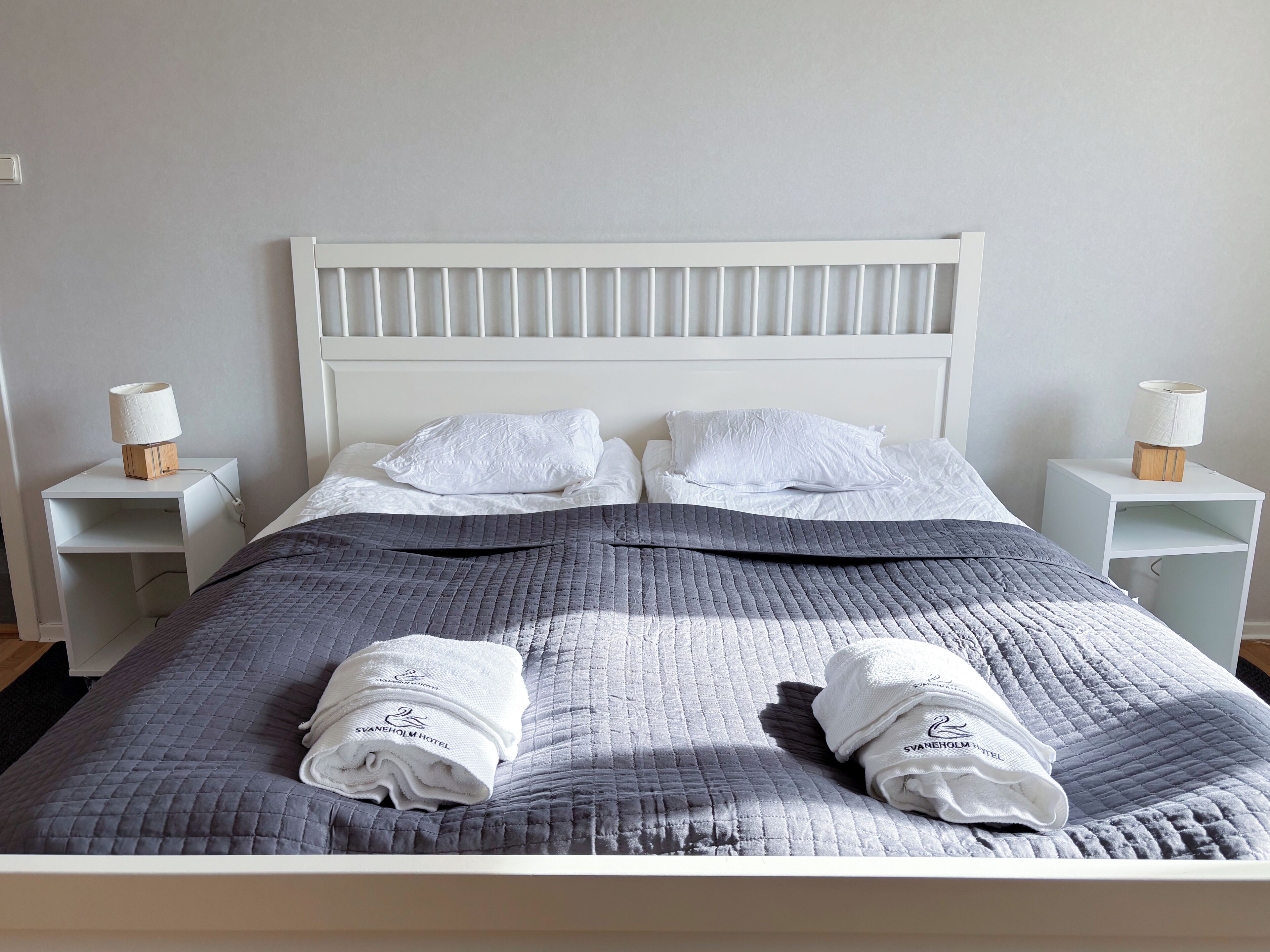 A neatly made bed with a grey quilt, two white pillows, and two folded towels with a hotel logo. There are two bedside tables with lamps and a white headboard.