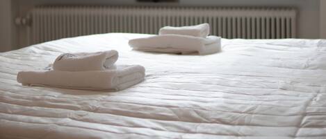 Una cama bien hecha con toallas dobladas, una ventana con vistas a edificios y un radiador debajo.