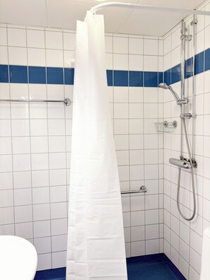 Un baño con paredes de azulejos blancos, una franja azul, una ducha con cabezal de ducha manual, mampara de vidrio y un inodoro blanco.