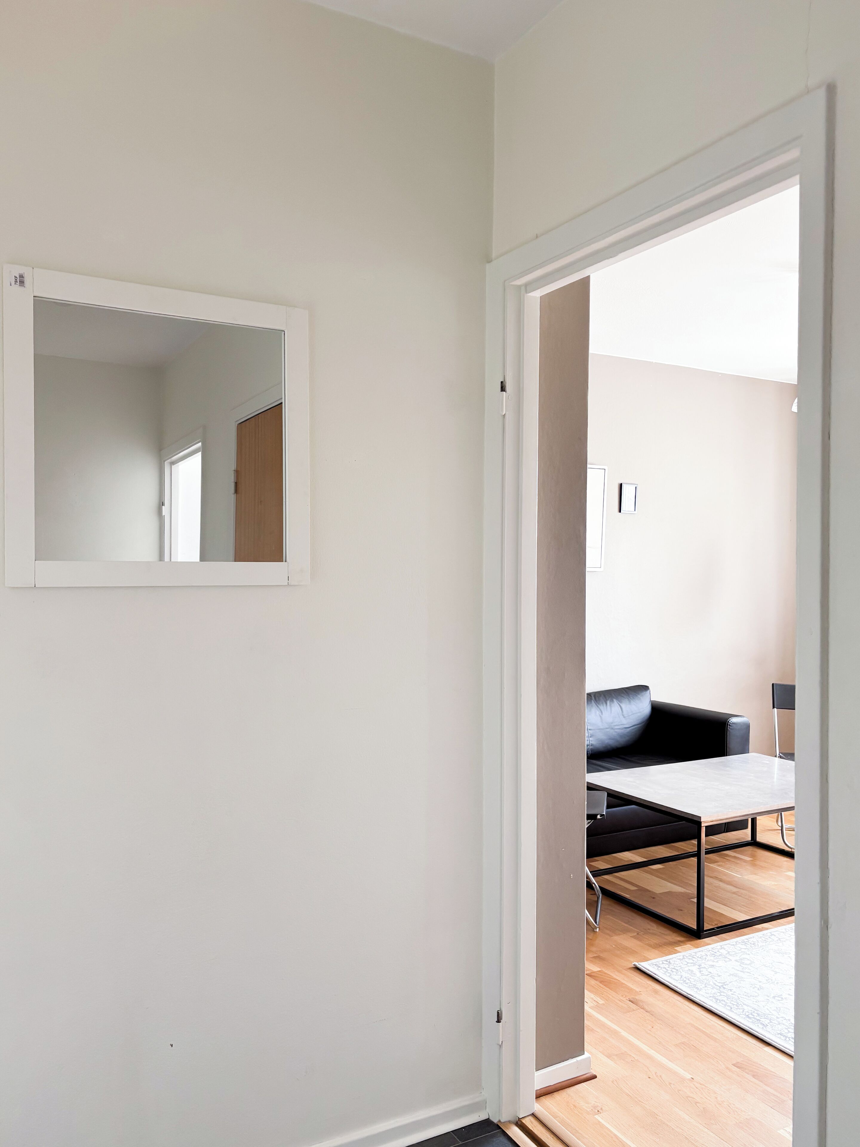 Una habitación con una puerta blanca, un espejo, un sofá negro, una mesita pequeña y un tapete estampado.