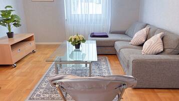 A modern living room with a glass coffee table, a grey sofa with decorative pillows, a small plant, and a folding chair.