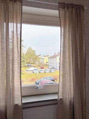 Una ventana con cortinas beige, vista de coches estacionados y edificios al fondo.