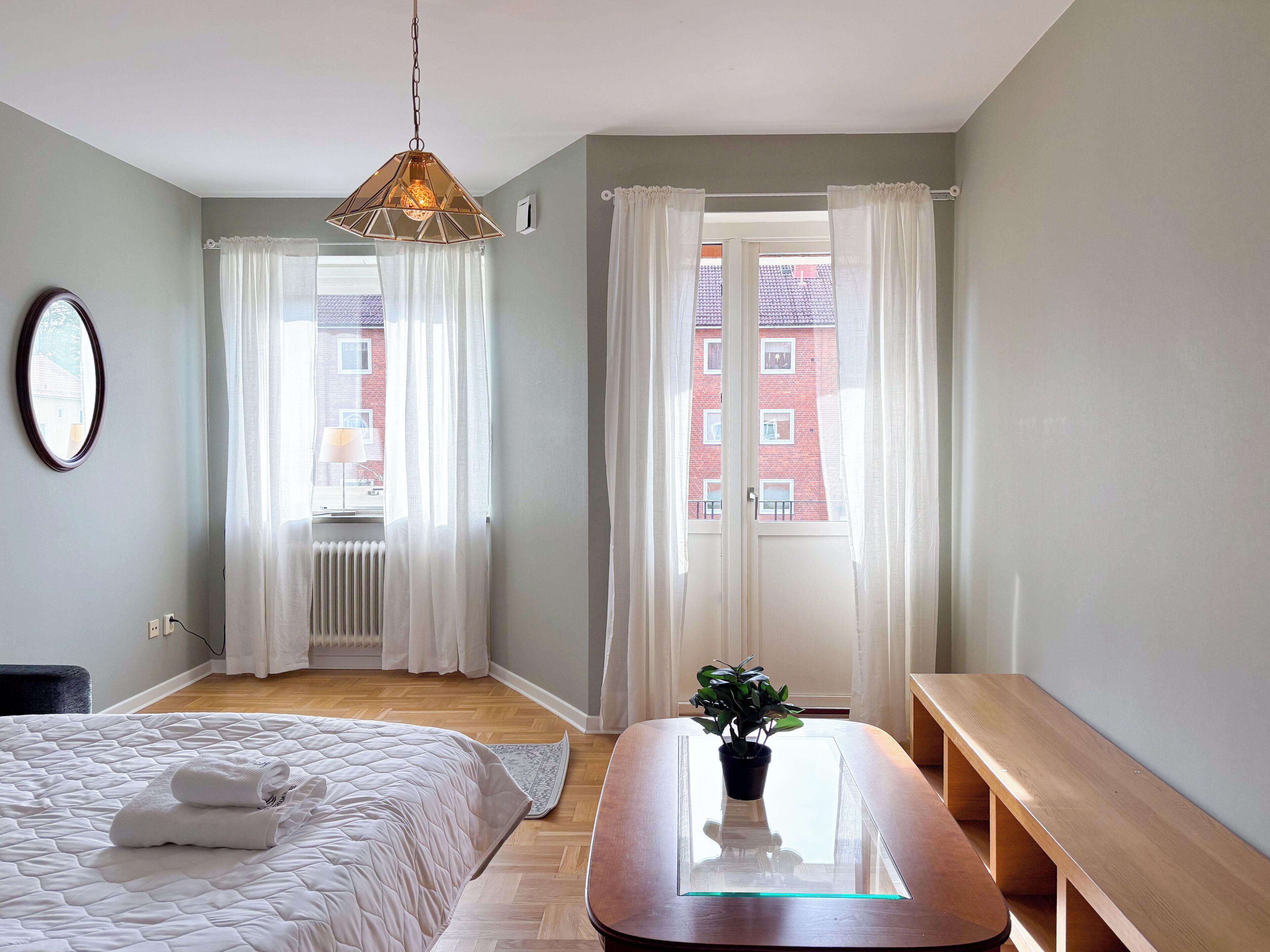 A bedroom with a bed, a bedside table with a plant, a hanging light fixture, and a view of a red building through the window.
