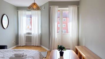 A bedroom with a bed, a bedside table with a plant, a hanging light fixture, and a view of a red building through the window.