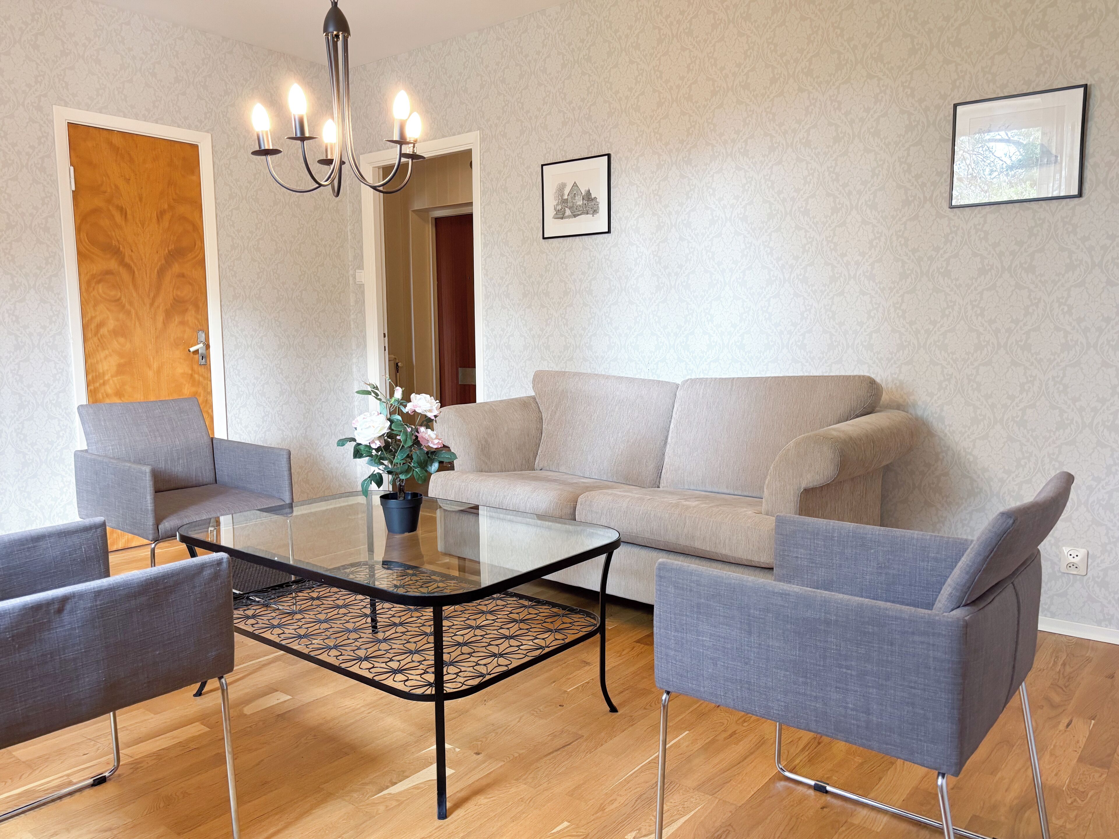 A living room with a sofa, two armchairs, a glass coffee table, a potted plant, and a chandelier.