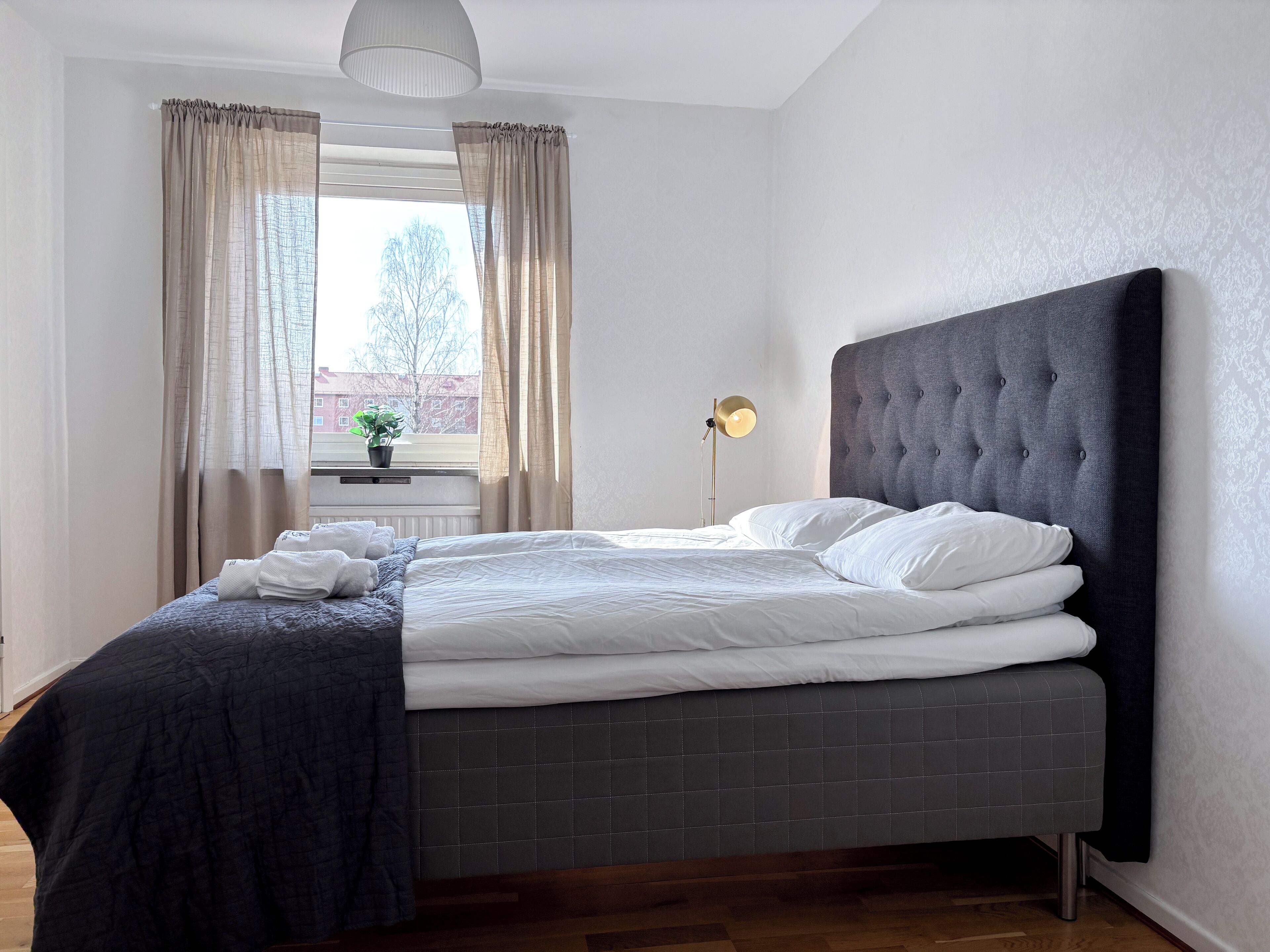 A neatly made bed with white linens and a dark headboard, a bedside lamp, and a window with curtains.