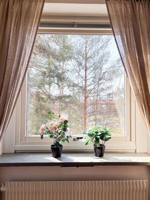 A window with two potted plants on a sill, curtains partially drawn, and a view of trees outside. - Säffle Suites - By Svaneholm Hotel (Saffle)